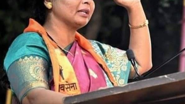Woman in a colorful sari raising her right hand while speaking at a podium, gesture of greeting or pledge.