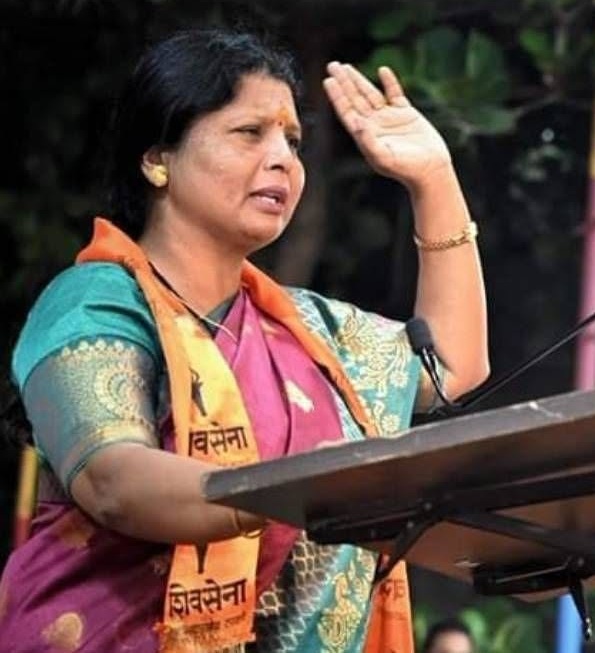 Woman in a colorful sari raising her right hand while speaking at a podium, gesture of greeting or pledge.