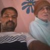Two people sit on a couch against a pink wall with a blue flower mural: a man in a striped shirt and an elderly woman with a brown headscarf.