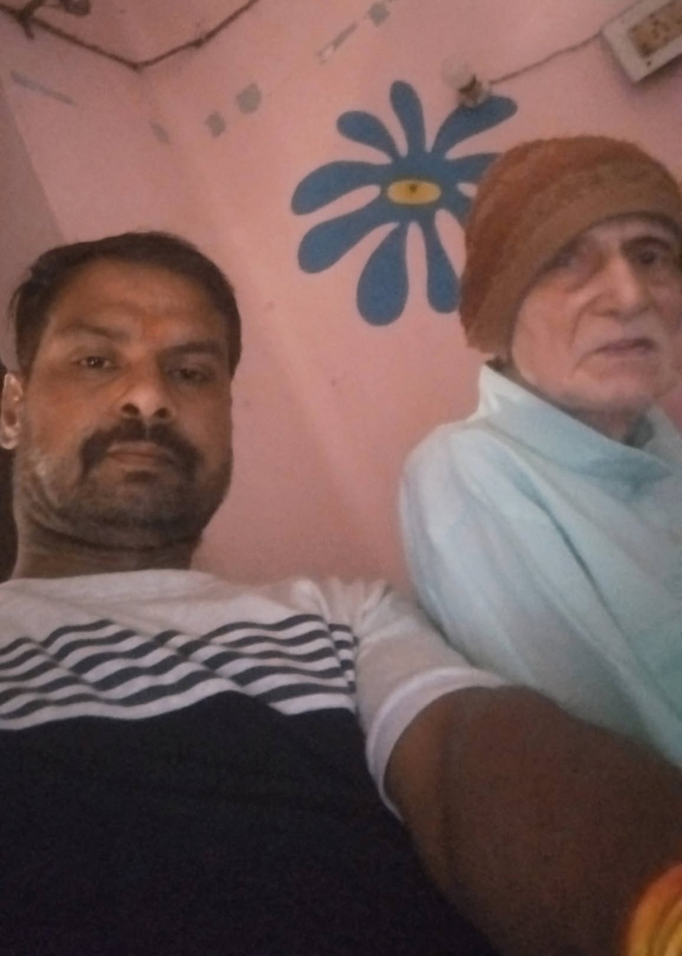 Two people sit on a couch against a pink wall with a blue flower mural: a man in a striped shirt and an elderly woman with a brown headscarf.