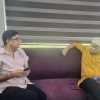 Two men sit on a purple sofa indoors, one in a pale pink shirt holding a smartphone while the other in an orange kurta and white cap rests a blue device in his hand, talking.