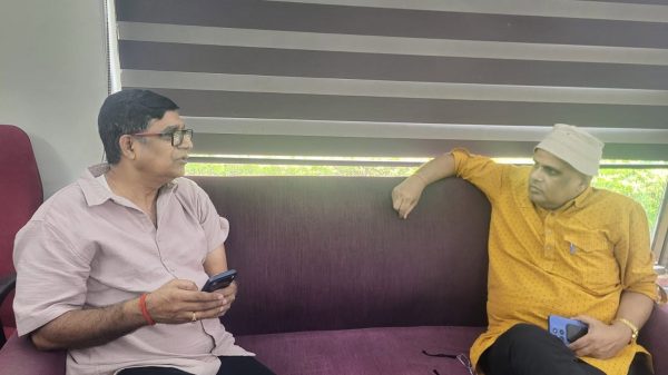 Two men sit on a purple sofa indoors, one in a pale pink shirt holding a smartphone while the other in an orange kurta and white cap rests a blue device in his hand, talking.