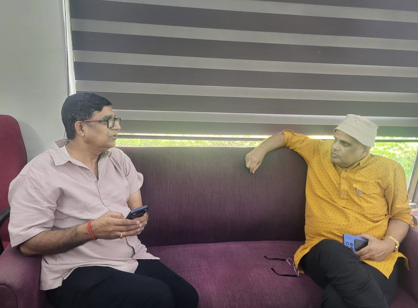 Two men sit on a purple sofa indoors, one in a pale pink shirt holding a smartphone while the other in an orange kurta and white cap rests a blue device in his hand, talking.