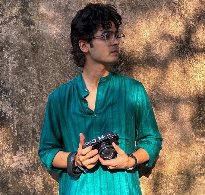 Young man in teal kurta holding a vintage camera, standing against a textured wall, wearing glasses and a watch.