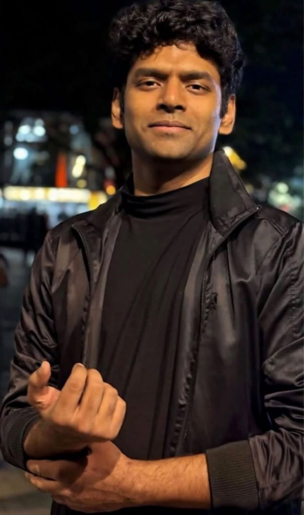 Young man in a black jacket standing outdoors at night with arms crossed or clasped in front of him.