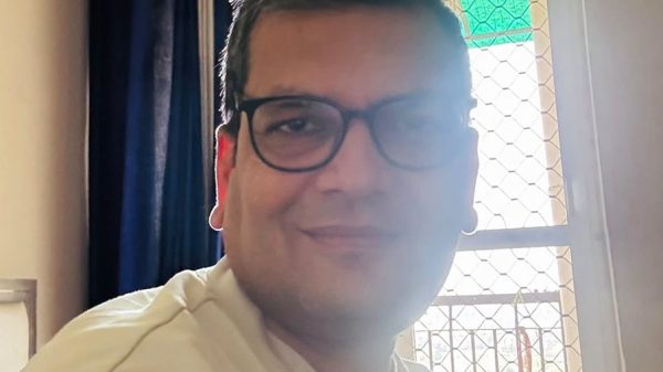 Smiling man with black-framed glasses wearing a light-colored shirt, indoors by a window with a mesh screen.