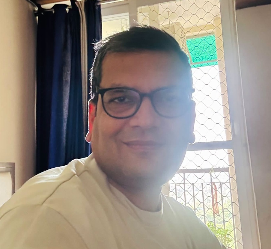 Smiling man with black-framed glasses wearing a light-colored shirt, indoors by a window with a mesh screen.