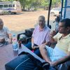 Three men sit around a wooden table outside, drinking from small cups; a water bottle and bags on the table with motorcycles and a dusty lot in the background.