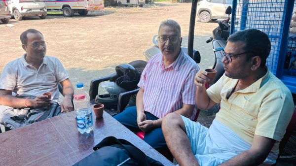 Three men sit around a wooden table outside, drinking from small cups; a water bottle and bags on the table with motorcycles and a dusty lot in the background.
