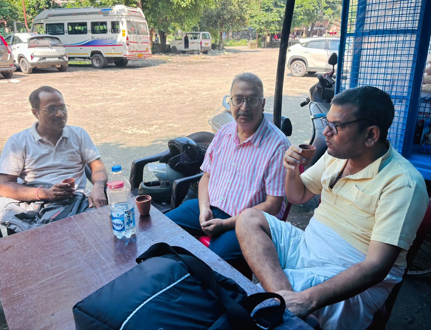 Three men sit around a wooden table outside, drinking from small cups; a water bottle and bags on the table with motorcycles and a dusty lot in the background.